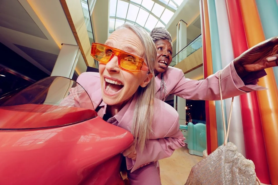 Two elderly women in pink suits ride a red toy car inside a shopping mall with colorful pillars and a glass roof, fun and cheerful.