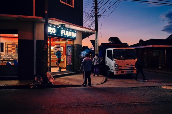 Man carries boxes into a store in a small town at dusk, with a delivery truck and other people visible on the street.
