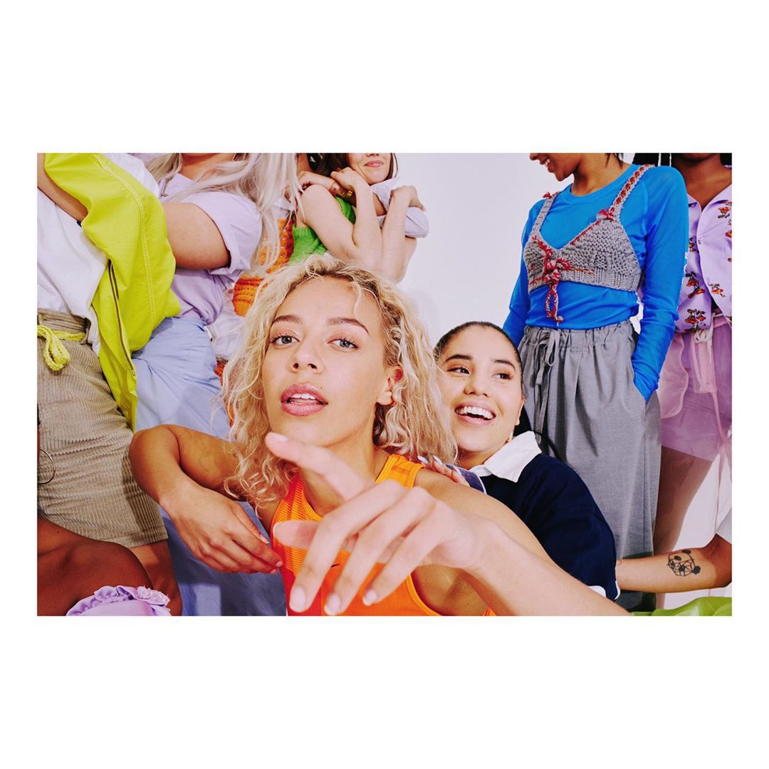 Diverse group of young adult women gather in a bright photo studio, wearing colorful clothes, posing, and smiling at the camera.