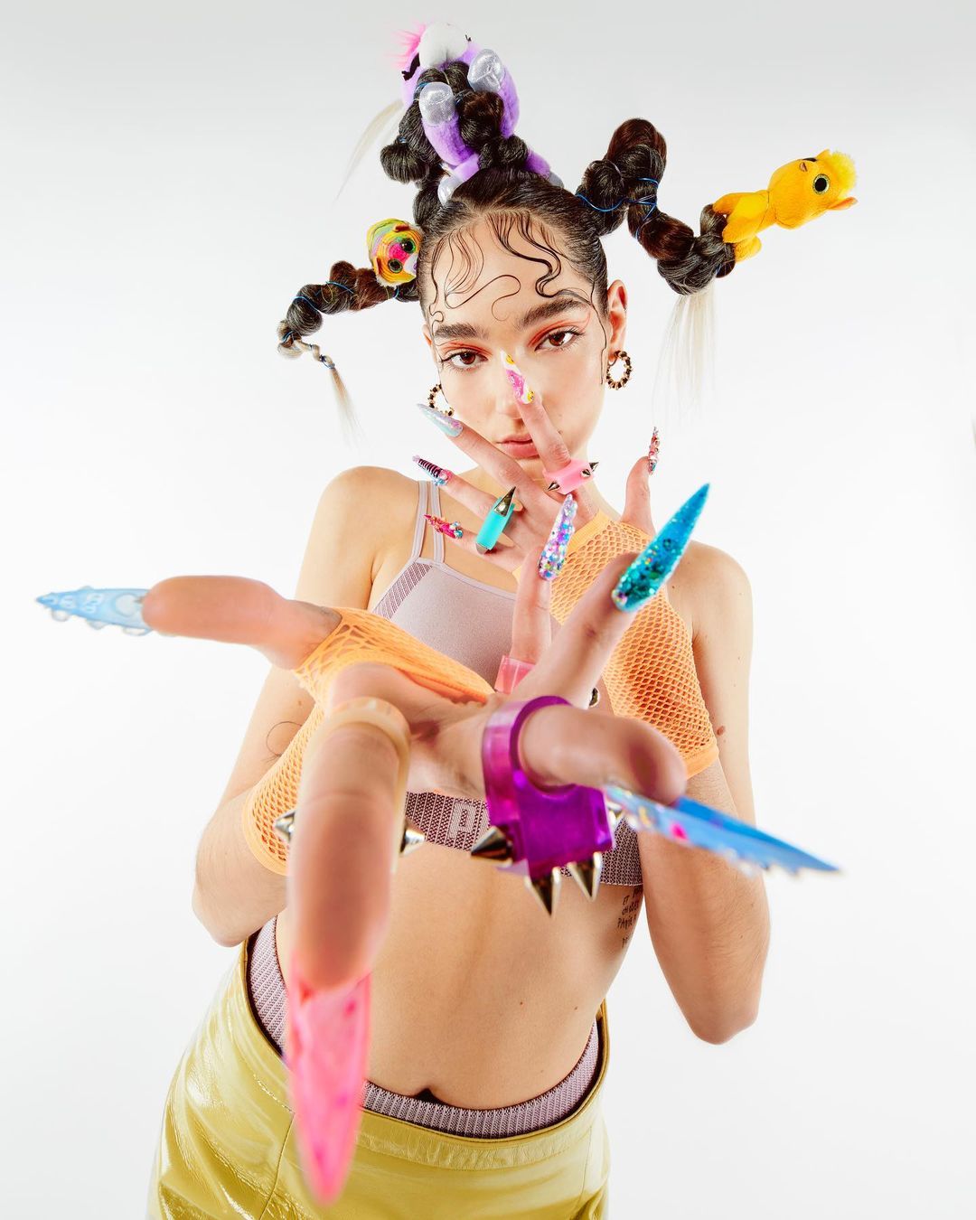 Woman with stylized hair and elaborate nail art poses in front of a white background, showcasing fashion and beauty elements.