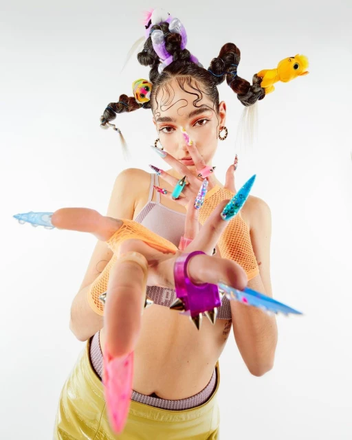 Woman with stylized hair and elaborate nail art poses in front of a white background, showcasing fashion and beauty elements.
