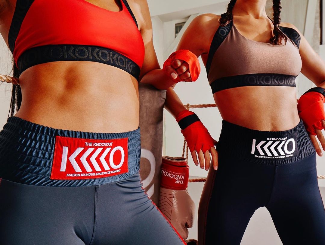 Two athletic women in sports bras and boxing-style shorts pose in a boxing ring with a punching bag. Athletic fitness fashion.