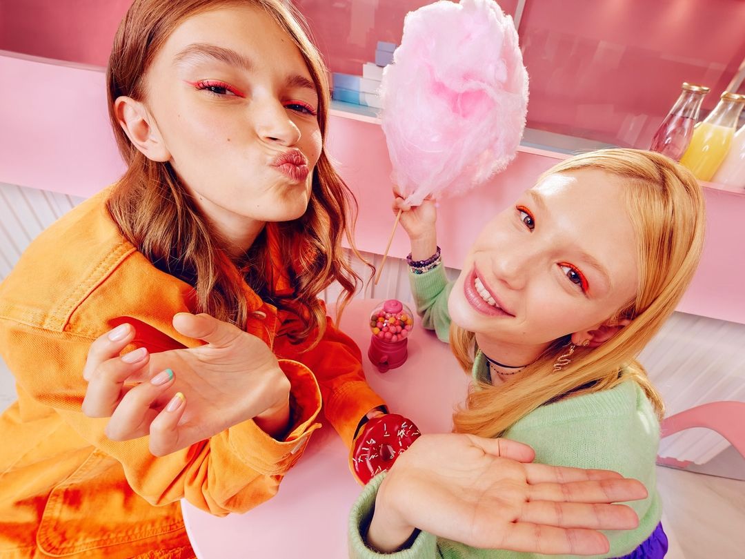 Two young women pose at a pink table with a donut, candy, and cotton candy, both wearing bright eye makeup and casual clothing.