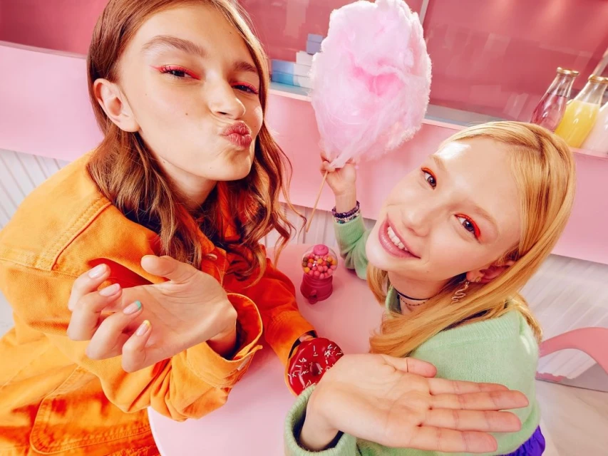 Two young women pose at a pink table with a donut, candy, and cotton candy, both wearing bright eye makeup and casual clothing.