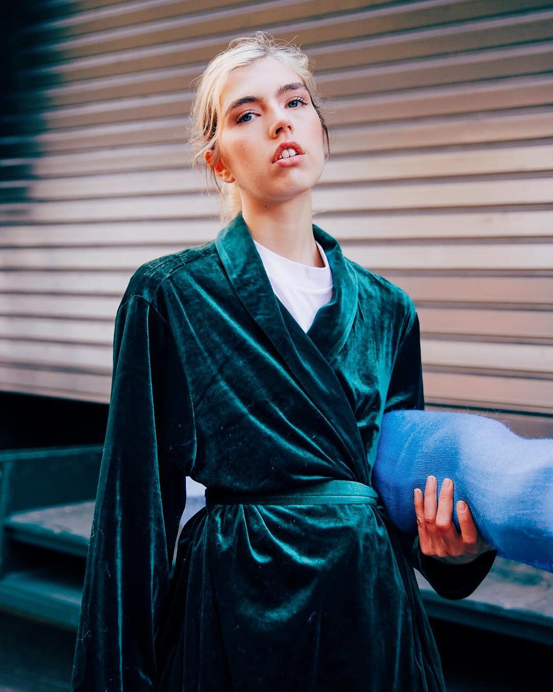 A blonde woman with fair skin stands outside wearing a green velvet robe over a white top, holding a light blue soft object.