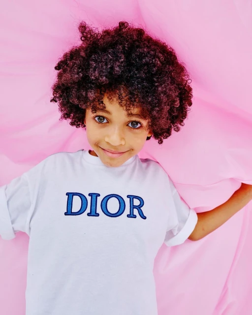 Close-up of a young child with curly hair and a Dior t-shirt, set against a vibrant pink backdrop. The child gazes directly at the camera.