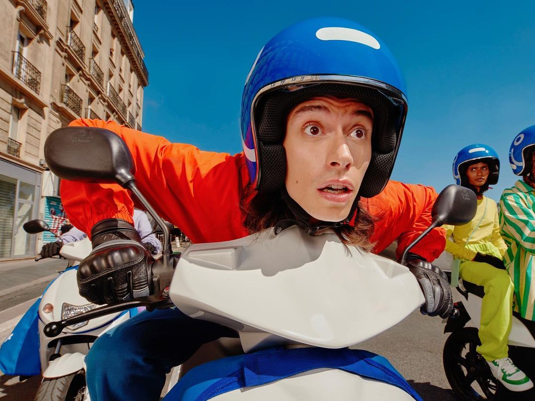 Delivery drivers on scooters in an urban setting with blue sky. The front driver looks surprised with an open mouth and wide eyes.