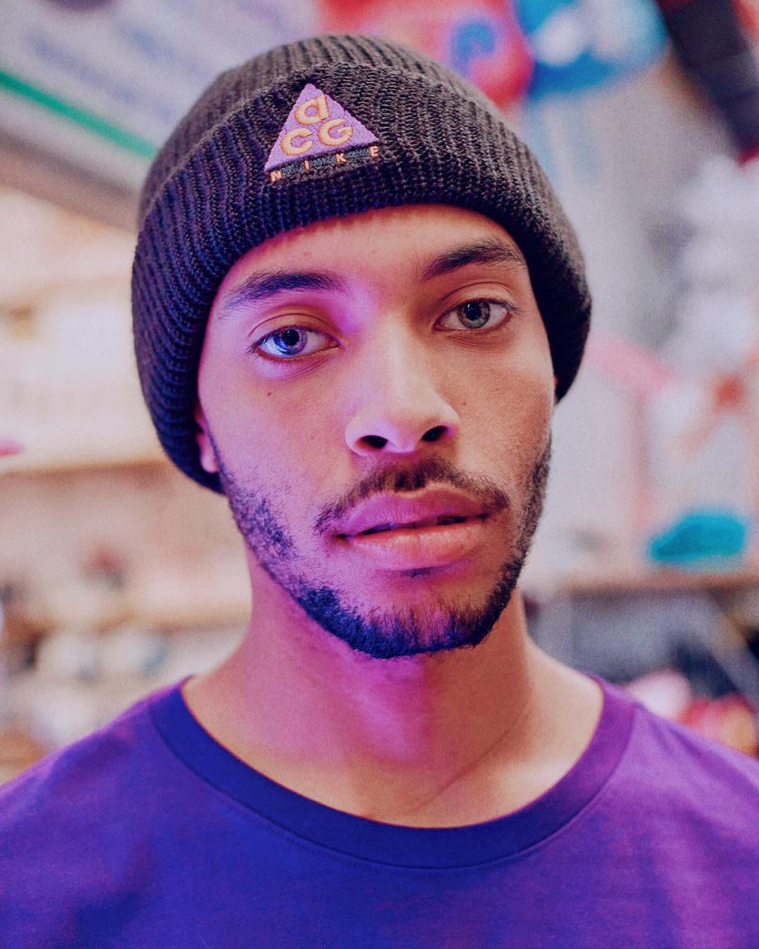Close-up studio shot of an adult male wearing a black beanie and purple t-shirt, looking directly at the camera, with soft lighting.