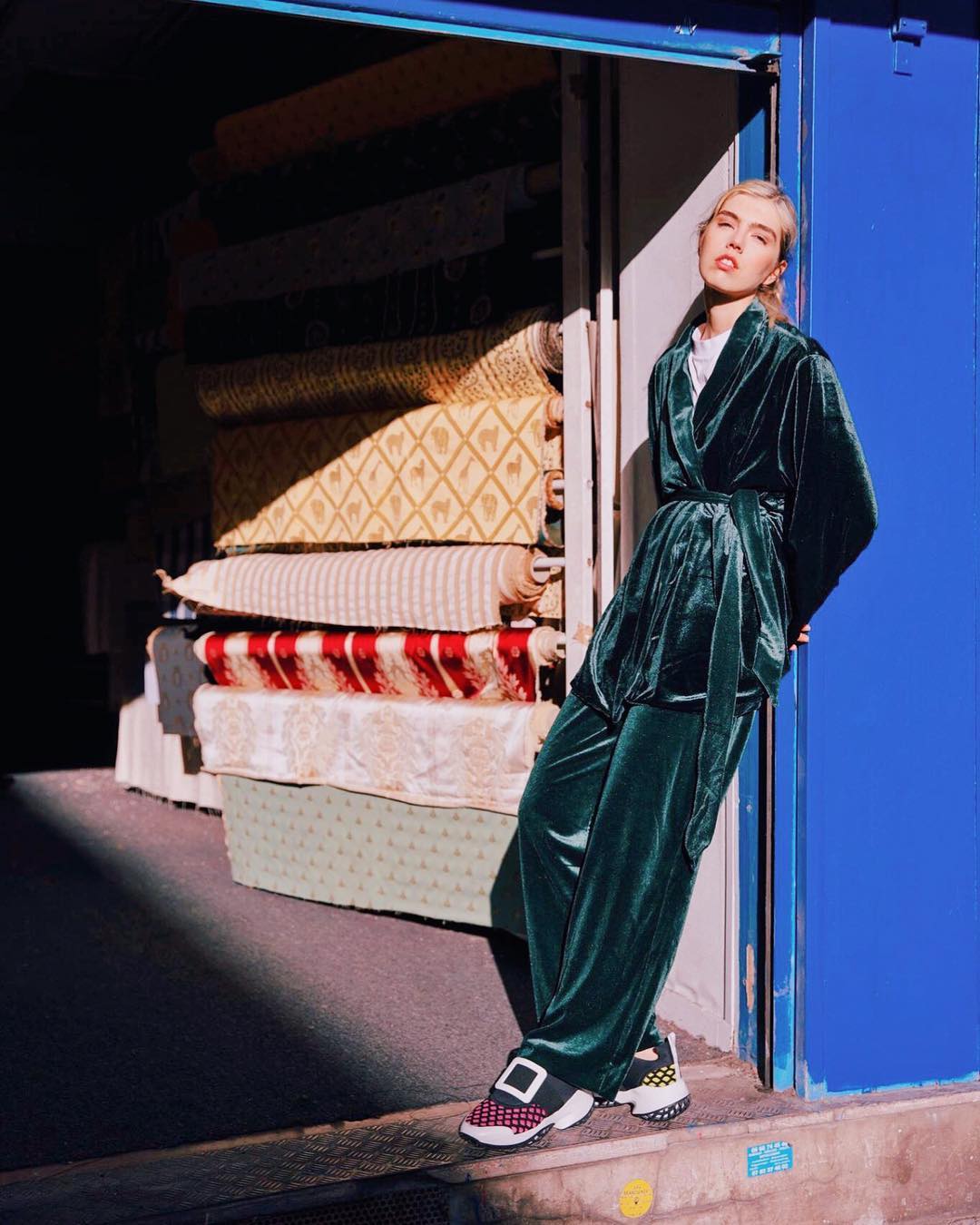 A fair-skinned woman in a green velvet suit poses in front of a blue doorway, next to a stack of patterned textiles outdoors.