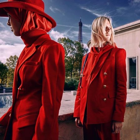 Two women in red suits pose near the Eiffel Tower. The woman on the left wears a red hat, while the other has blonde hair.