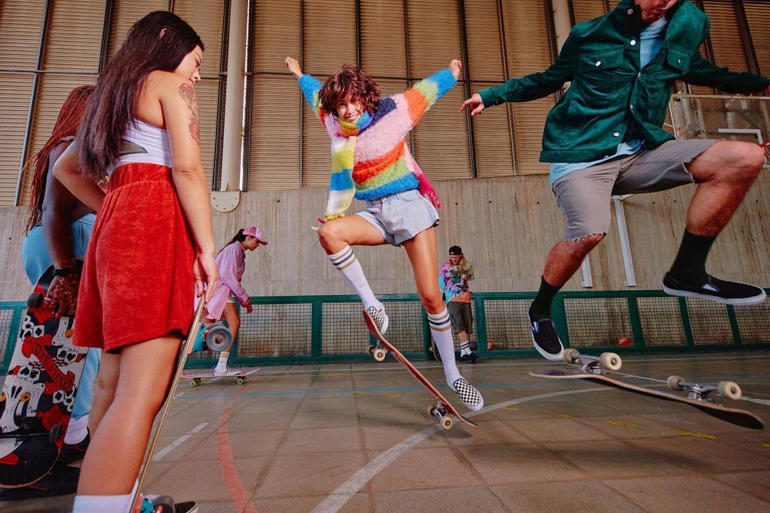 Group of young adults skateboarding in an indoor skatepark, mid-air jump, wearing casual trendy clothing, skateboards, athletic socks, and sneakers.