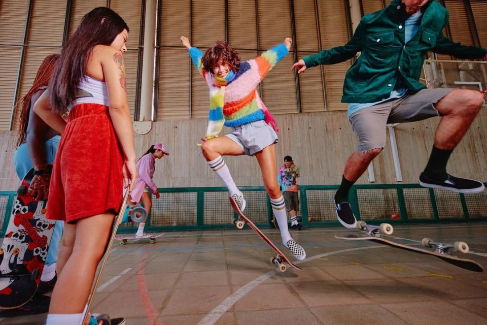 Group of young adults skateboarding in an indoor skatepark, mid-air jump, wearing casual trendy clothing, skateboards, athletic socks, and sneakers.