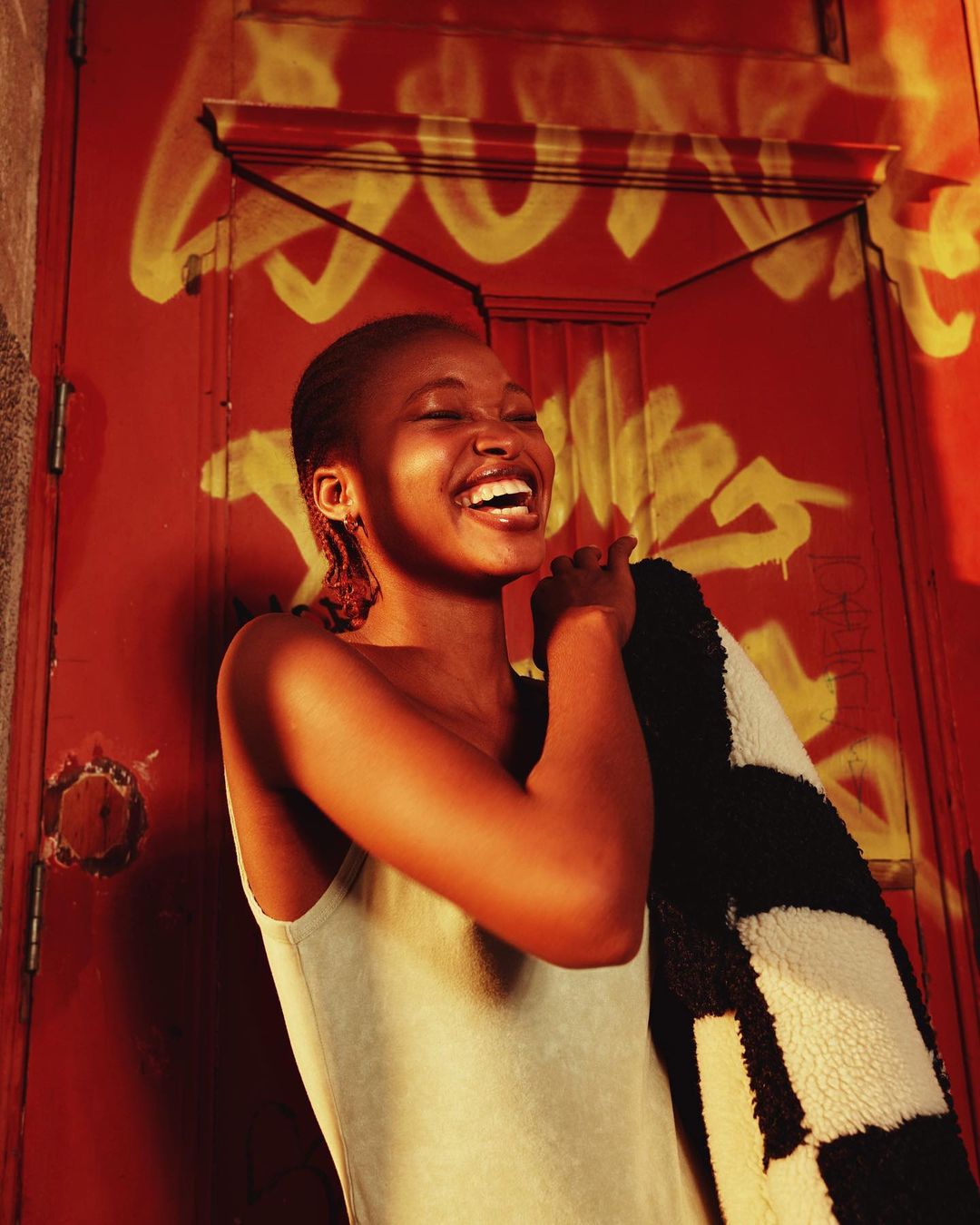 A smiling African American woman with an afro wearing a tank top and holding a black and white jacket in front of a red door with graffiti.