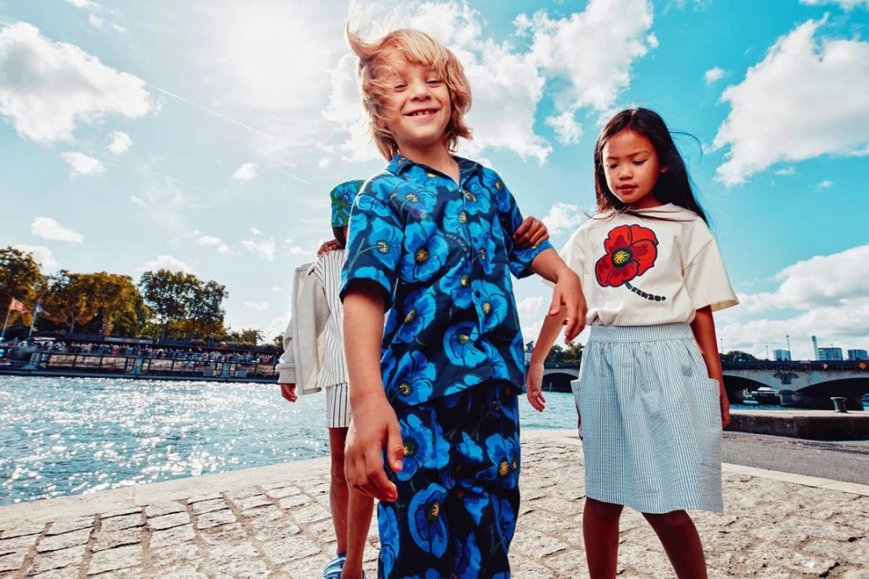 A group of children wearing floral and striped outfits are standing on a stone walkway by a river under a partly cloudy sky.