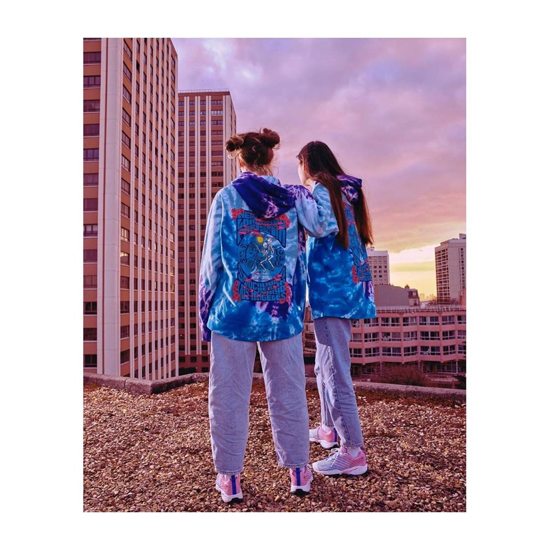 Two women stand on a rooftop in tie-dye hoodies and jeans against a city backdrop with a sunset sky, showcasing casual urban fashion.
