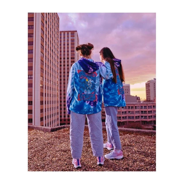 Two women stand on a rooftop in tie-dye hoodies and jeans against a city backdrop with a sunset sky, showcasing casual urban fashion.