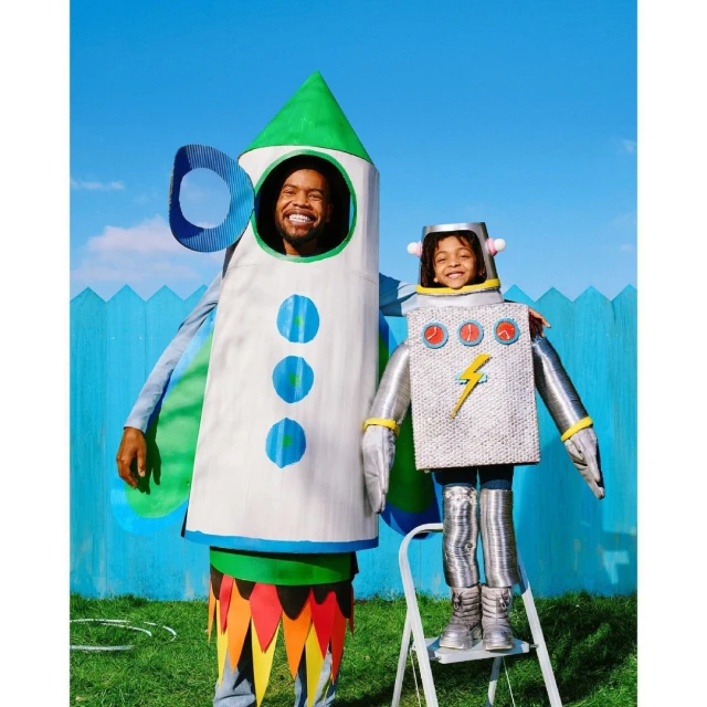 Adult male and child outdoors dressed in homemade rocket and robot costumes with a blue sky and green grass background.