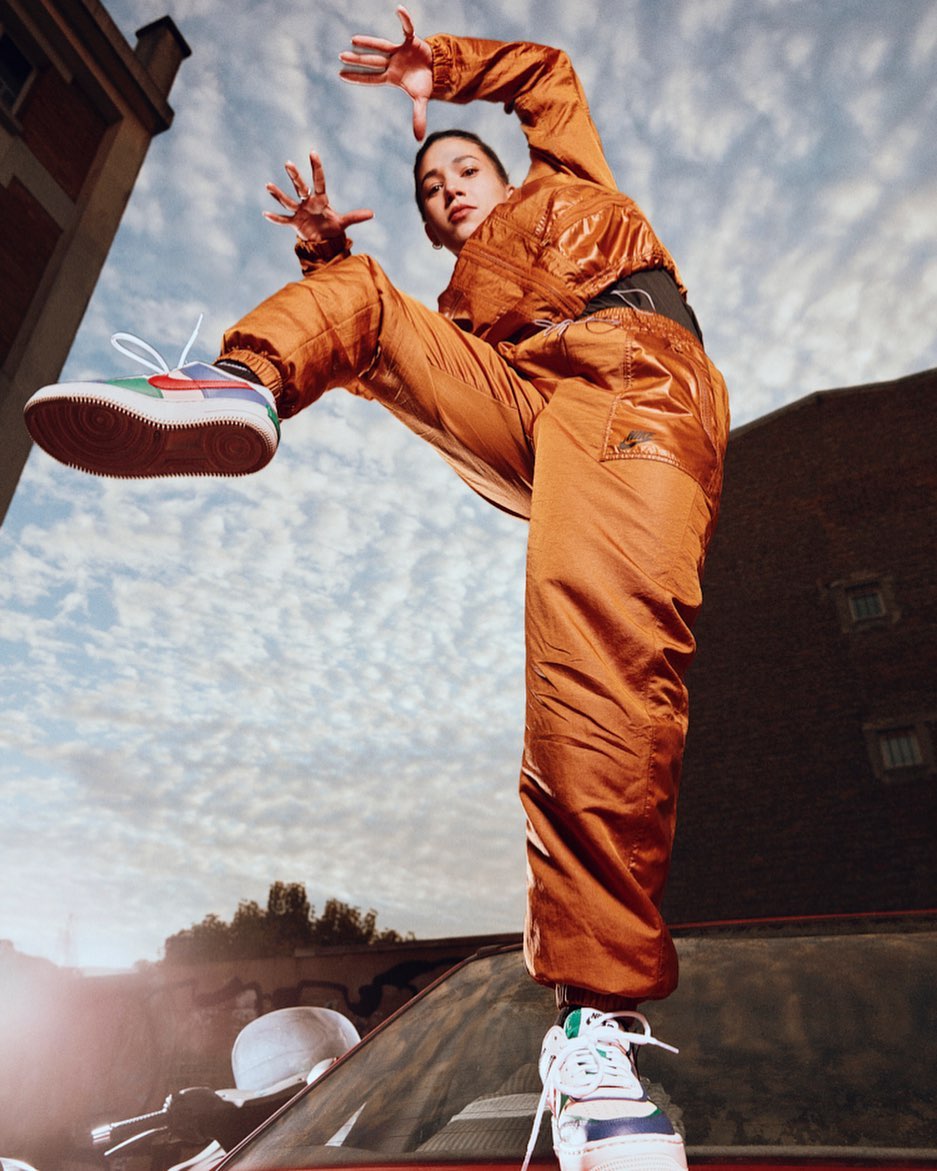 A young Asian woman in a bright orange outfit balances on a car, striking a pose against a cloudy sky and brick buildings.