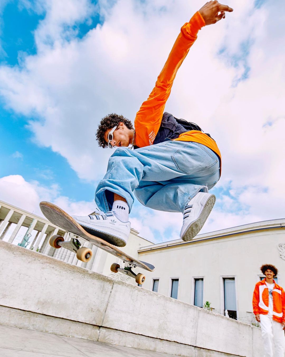 Young adult male performs a skateboard trick in mid-air wearing a bright orange shirt, loose jeans, and white sneakers against a bright sky.