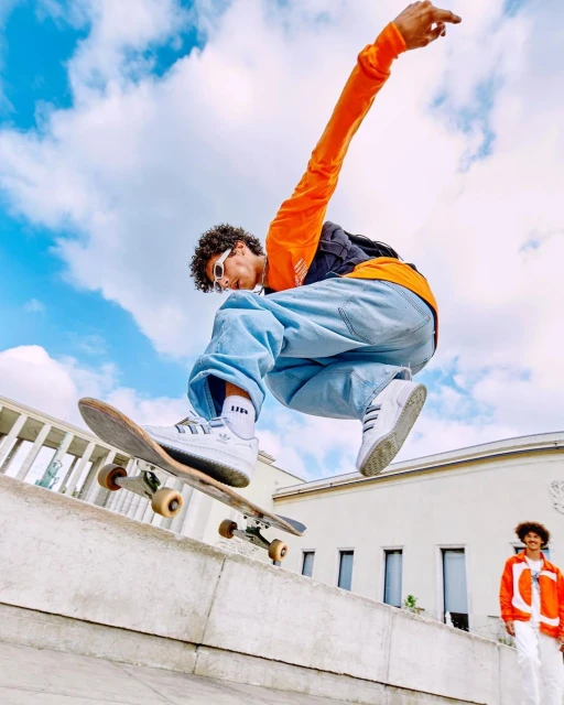 Young adult male performs a skateboard trick in mid-air wearing a bright orange shirt, loose jeans, and white sneakers against a bright sky.