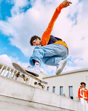 Young adult male performs a skateboard trick in mid-air wearing a bright orange shirt, loose jeans, and white sneakers against a bright sky.