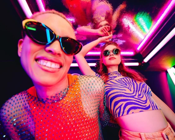 Two stylish people pose in vibrant lighting wearing sunglasses and colorful clothing. Fun, fashionable portrait with dynamic energy. 