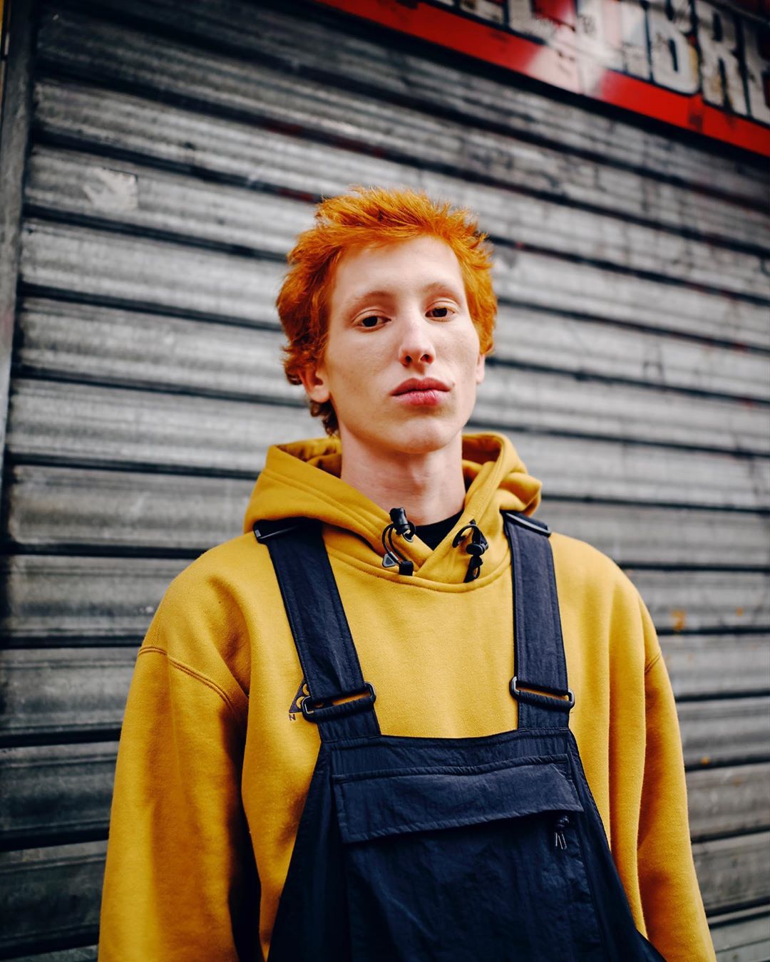 Young adult with ginger hair wearing overalls and a yellow hoodie posing in front of a metal shutter in an urban setting.