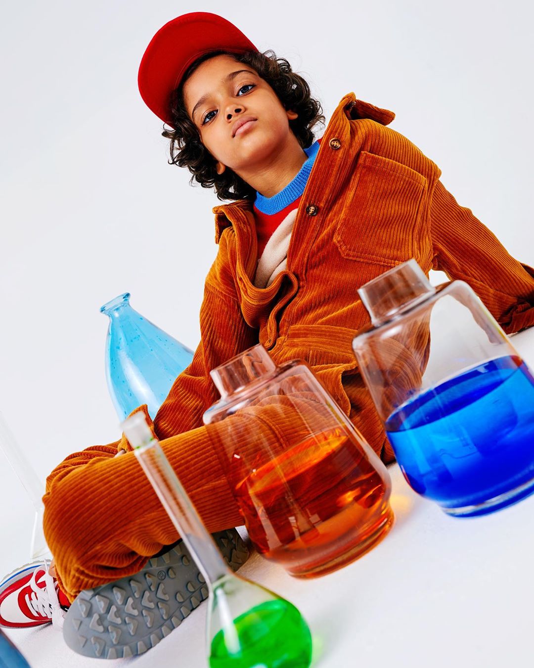 Young child in a brown corduroy outfit and red cap sitting surrounded by colorful glass laboratory beakers and bottles on white backdrop.