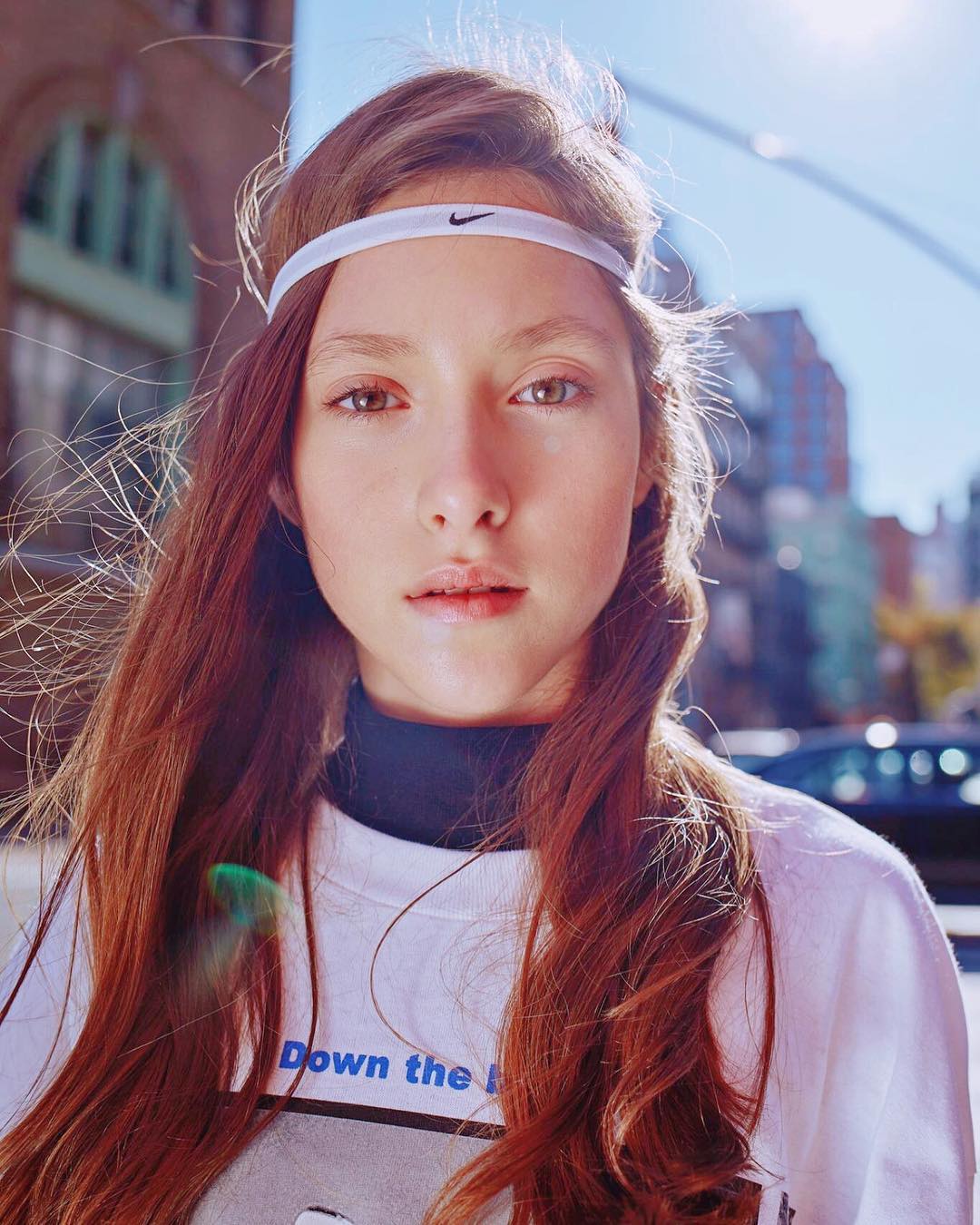 Close-up of a young woman with auburn hair and a Nike headband standing in a city street with buildings in the background on a sunny day.