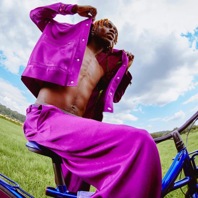 A shirtless man in a magenta suit is riding a bike in a grassy field under a sky with fluffy clouds in a fashion-focused image.