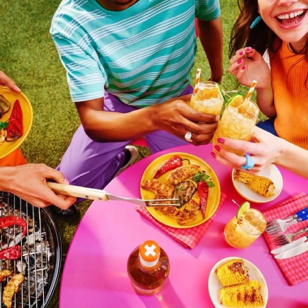 Overhead shot of friends grilling food and enjoying cocktails outside on a sunny day with grilled corn, peppers, and chicken.