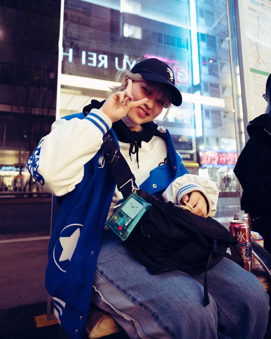 A young adult with a baseball cap and varsity jacket sits outside, making a peace sign. A clear BMO bag is slung across their body.