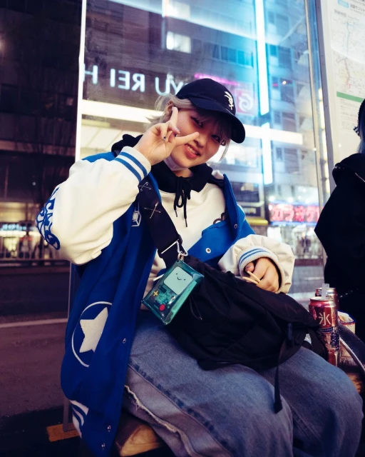 A young adult with a baseball cap and varsity jacket sits outside, making a peace sign. A clear BMO bag is slung across their body.