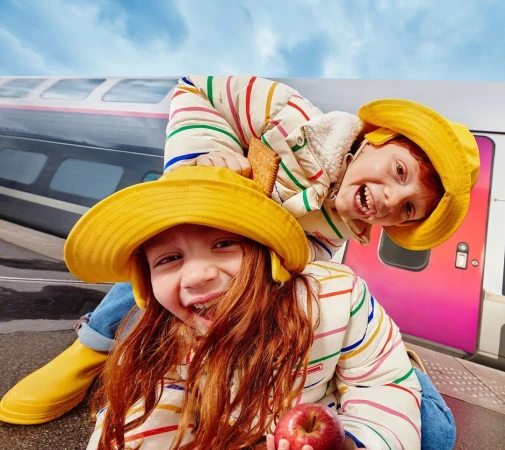 Two red-haired children in yellow hats and striped jackets pose in front of a train. One child carries a snack, the other holds a red apple.