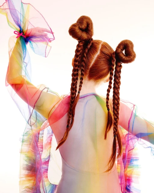 A child model with red hair in pigtails, wearing a sheer rainbow dress, poses in a studio with a white backdrop, showcasing a whimsical style.