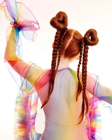 A child model with red hair in pigtails, wearing a sheer rainbow dress, poses in a studio with a white backdrop, showcasing a whimsical style.