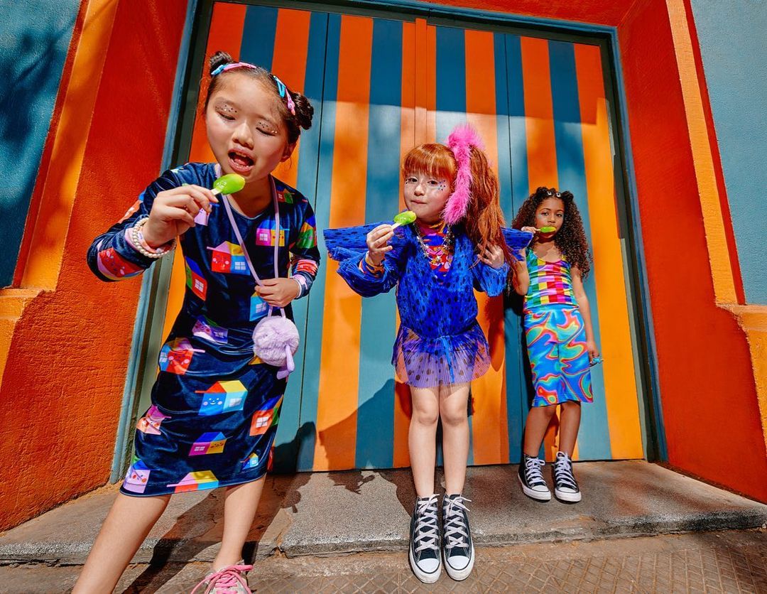 Three young girls in vibrant patterned clothing stand against a bold, vertically striped orange and blue wall, each enjoying a lollipop.