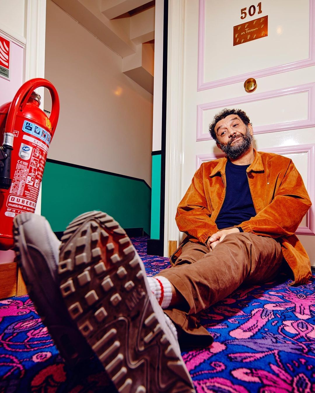 Man in brown jacket sits with legs out, Nike shoe sole facing camera on patterned carpet in corridor, next to fire extinguisher.