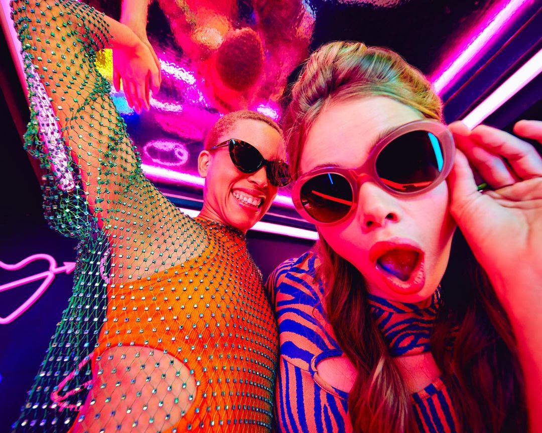 Two people in colorful outfits and sunglasses pose indoors with neon lighting, creating a vibrant and energetic visual.