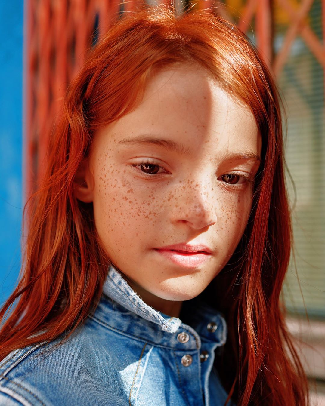 Close-up portrait of a red-haired girl with freckles wearing a denim jacket, captured in soft, natural light against a textured background.