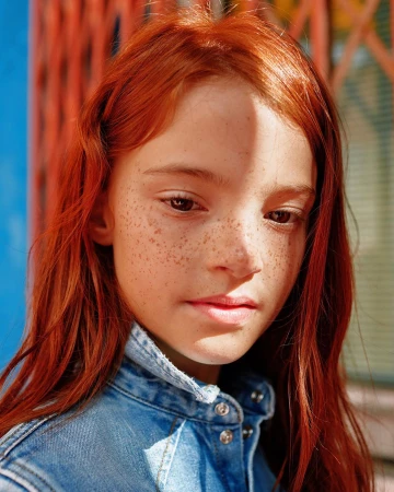 Close-up portrait of a red-haired girl with freckles wearing a denim jacket, captured in soft, natural light against a textured background.