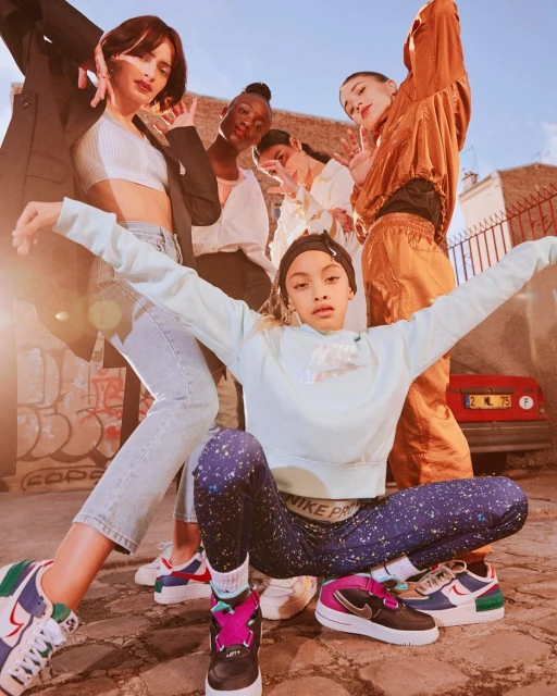 Group of six young women in an urban setting, wearing athletic and casual clothing and sneakers, posing and interacting in sunlight.