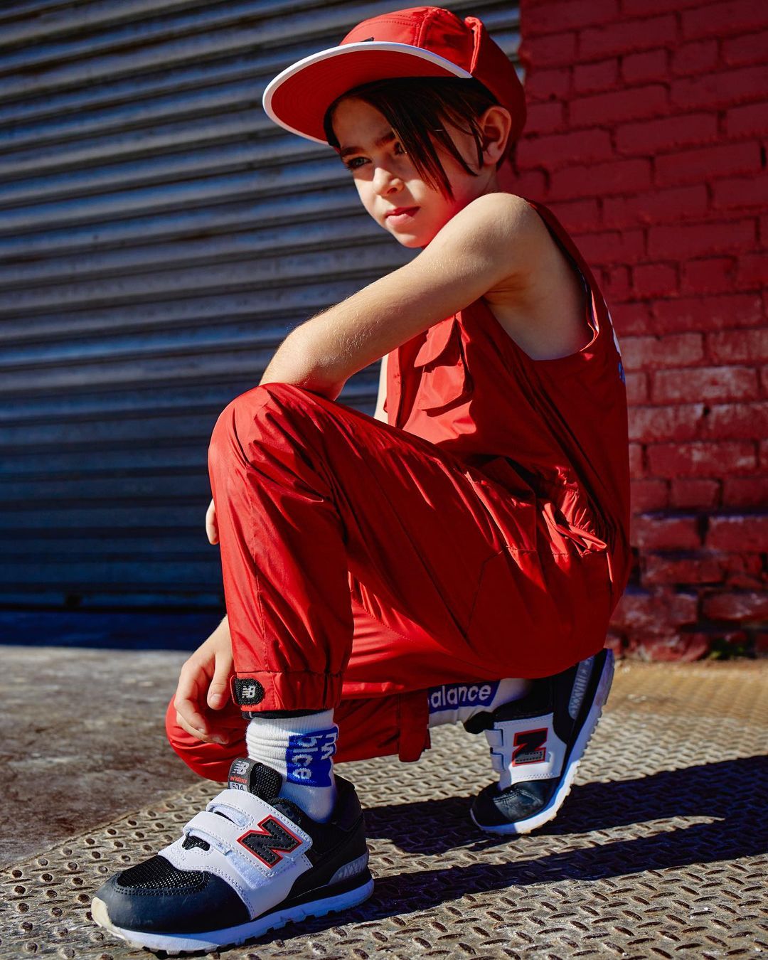 Child model in red outfit poses outdoors against corrugated metal and brick wall. Features New Balance shoes, cap, and branded socks.