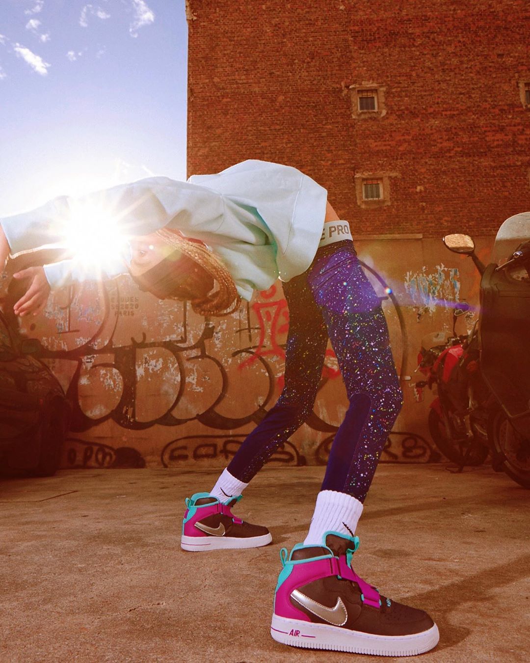 Young adult doing a backbend in athletic wear and bright-colored athletic shoes, in an urban setting with graffiti and a brick building.