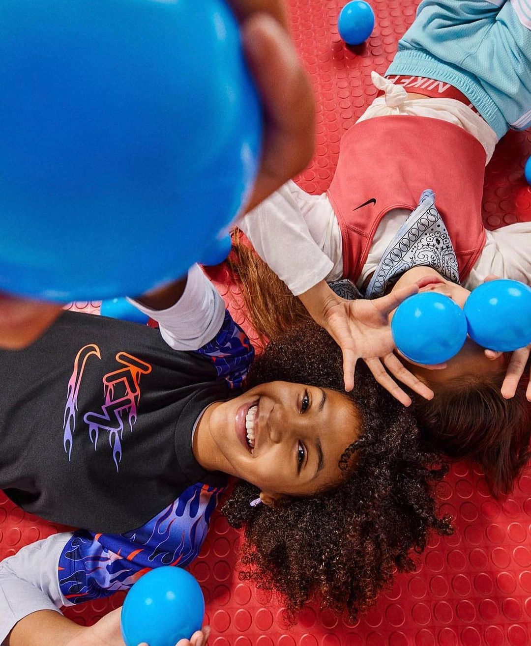 Two smiling kids playing with blue balls on a red polka dot surface, wearing colorful clothing, photographed from a high angle.