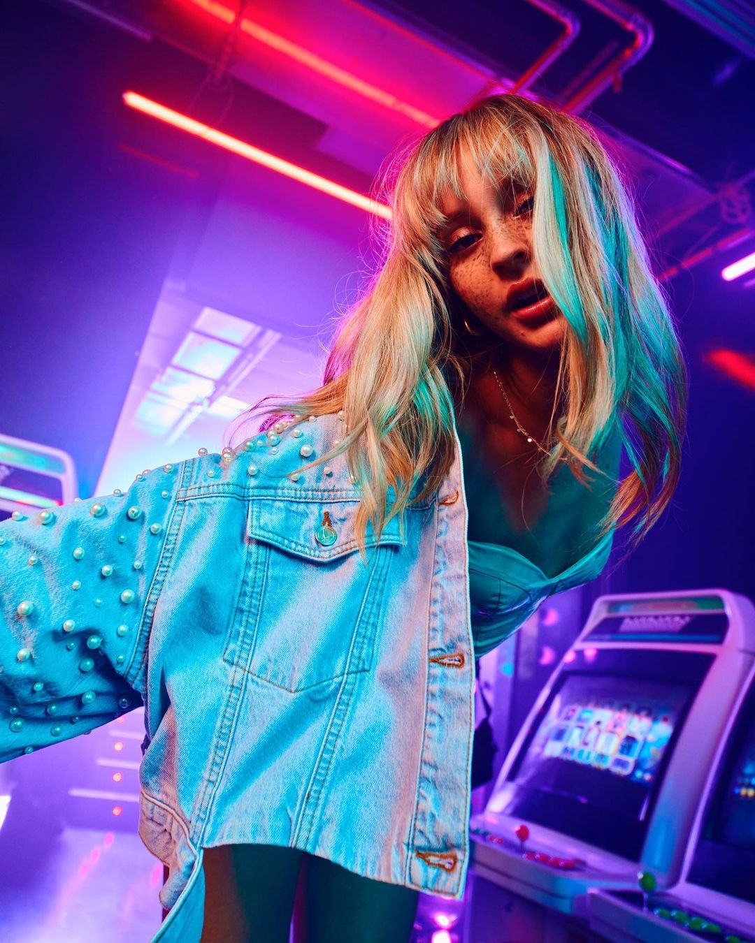 Blonde woman in denim jacket with pearl embellishments stands inside an arcade with bright neon lighting and gaming machines.