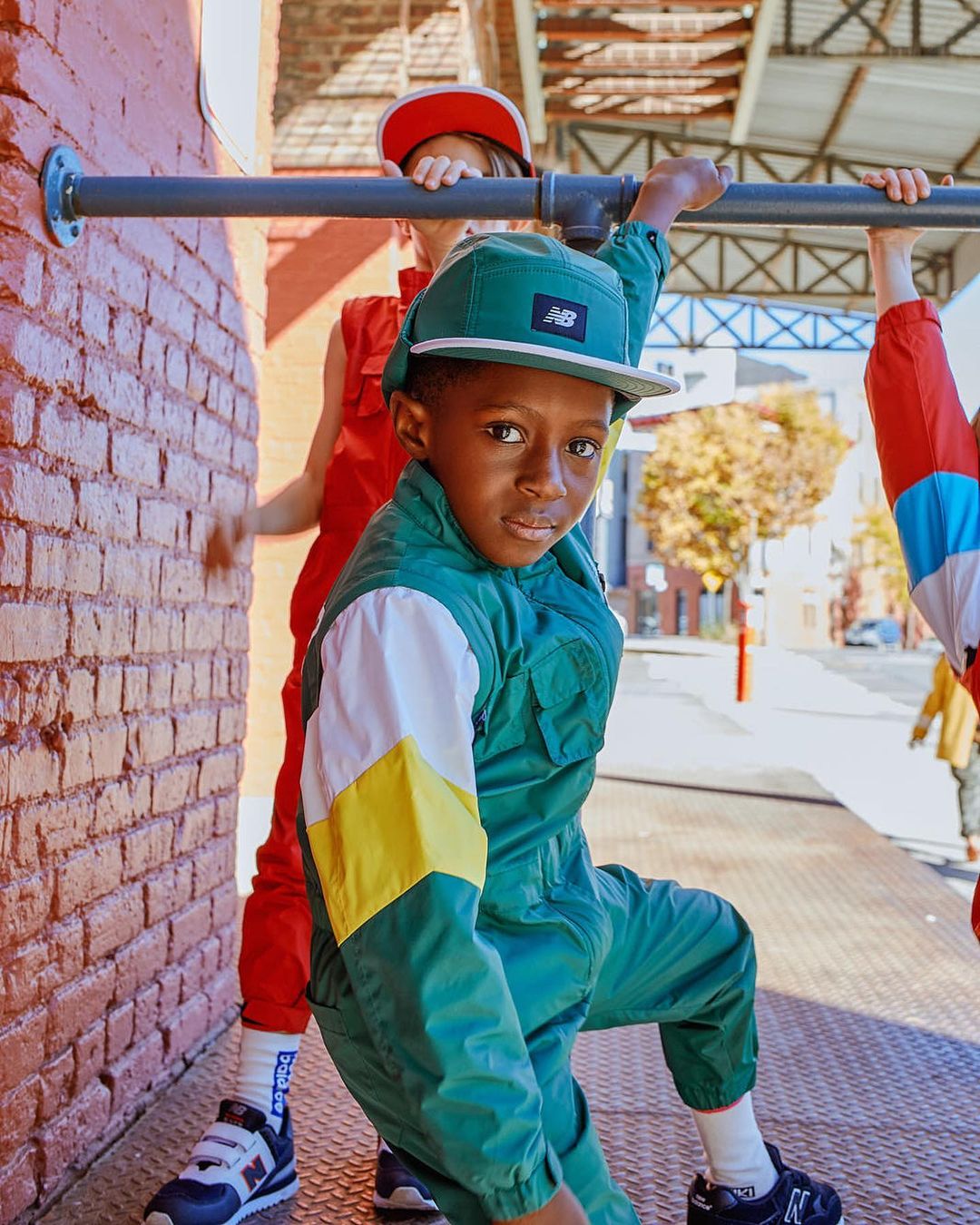 Child in retro-style green jumpsuit and matching cap poses with other kids in red jumpsuits by a brick wall outdoors in daytime.