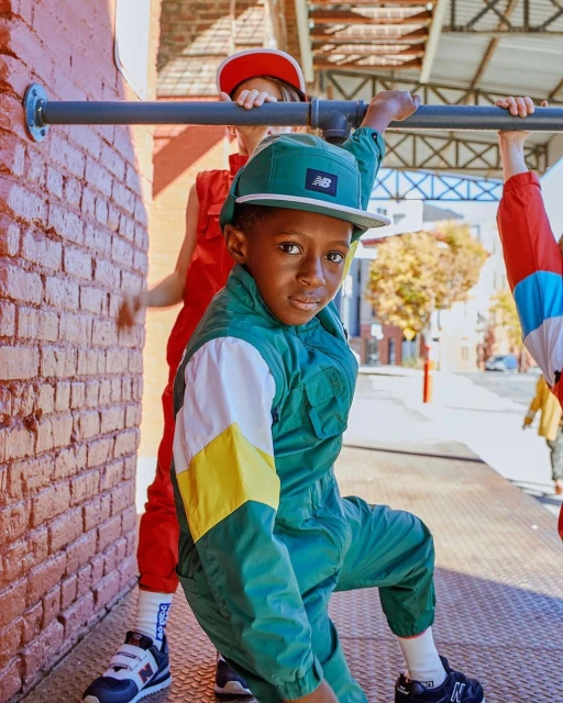 Child in retro-style green jumpsuit and matching cap poses with other kids in red jumpsuits by a brick wall outdoors in daytime.