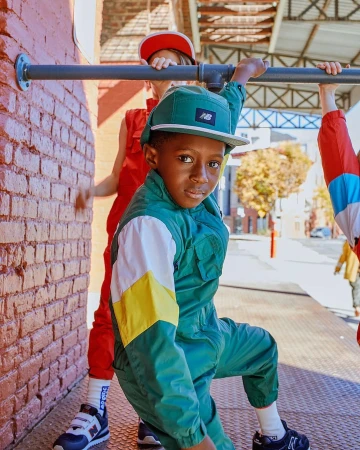 Child in retro-style green jumpsuit and matching cap poses with other kids in red jumpsuits by a brick wall outdoors in daytime.
