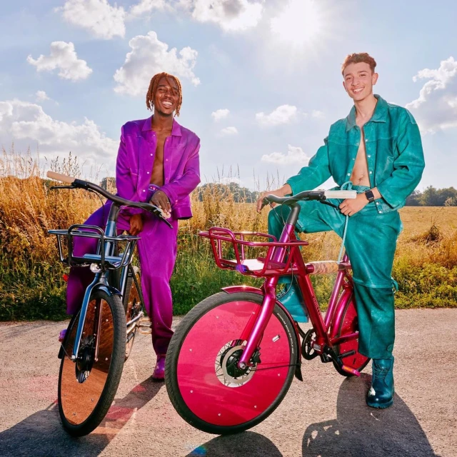 Two people pose outdoors with custom-designed bikes. One is purple and the other is teal, with fields and blue sky in the background.